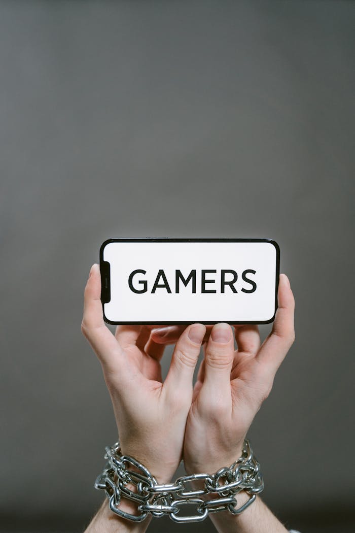 Person's hands in chains holding a smartphone displaying the word 'GAMERS' on a gray background.